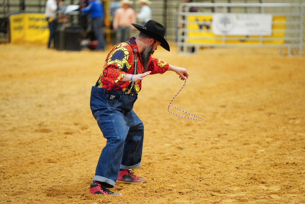 A rodeo clown wearing a red shirt and denim overalls twirling a small rope.