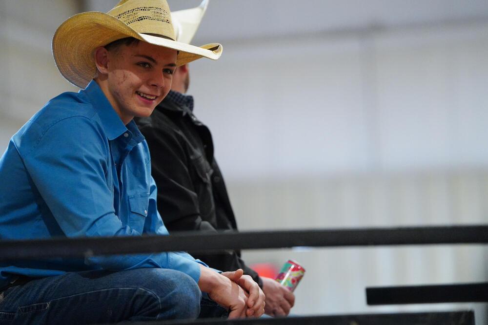 A teenage cowboy wearing a blue shirt and cowboy hat smiling at the camera while seated on a fence. 