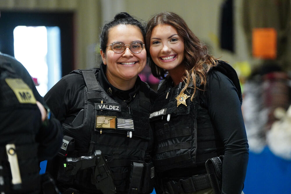 2 female corrections officers wearing black police vests smiling at the camera.