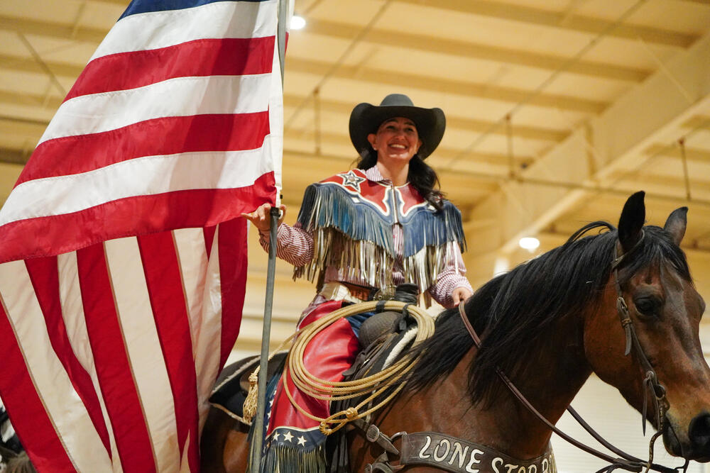 A cowgirl wearing a red, white and blue fringed jacket on horseback carrying the American Flag.