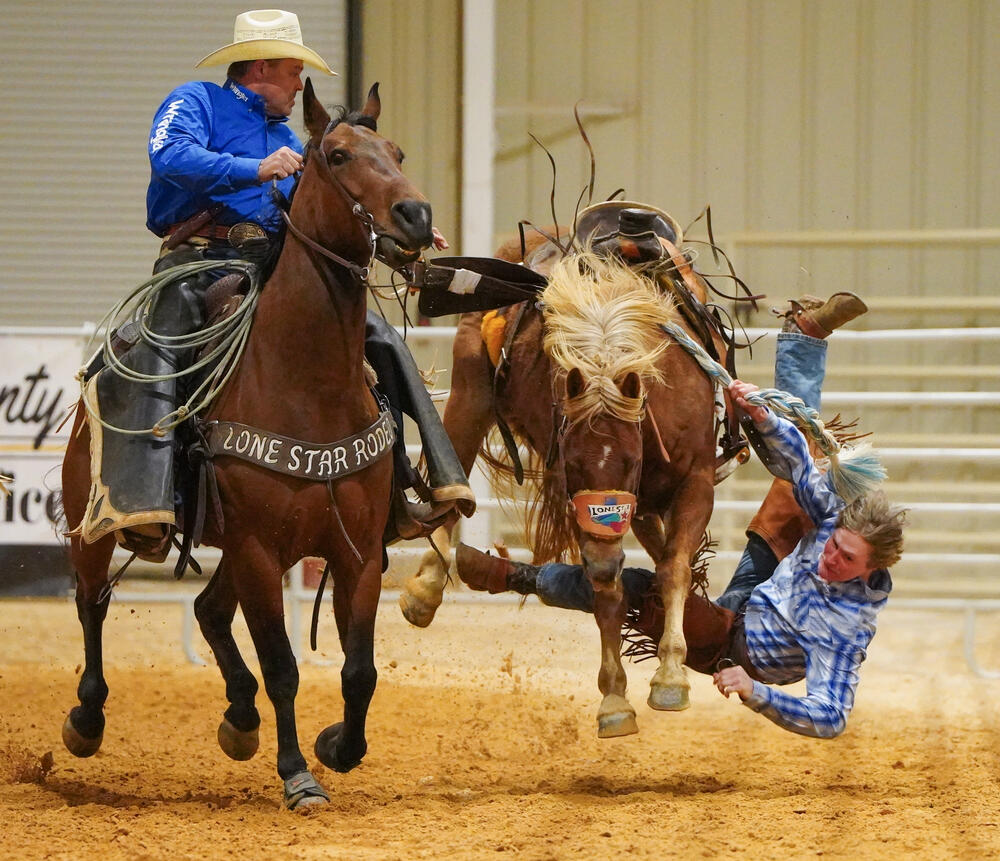 A cowboy in midair falling from a bucking horse while another cowboy on horseback attempting to corral the bucking horse. 