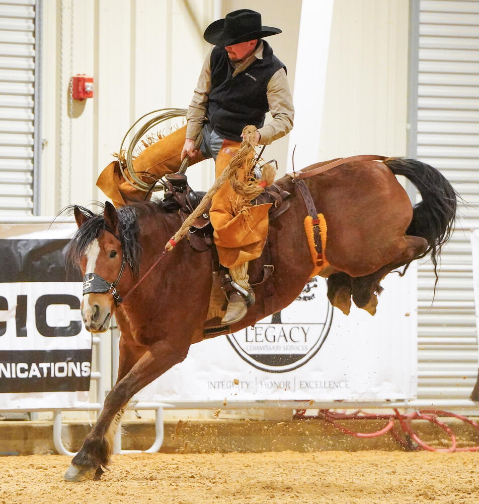 A cowboy wearing a black vest, tan shirt and orange chaps, holding onto the reigns of a bucking bronco will in the air. 