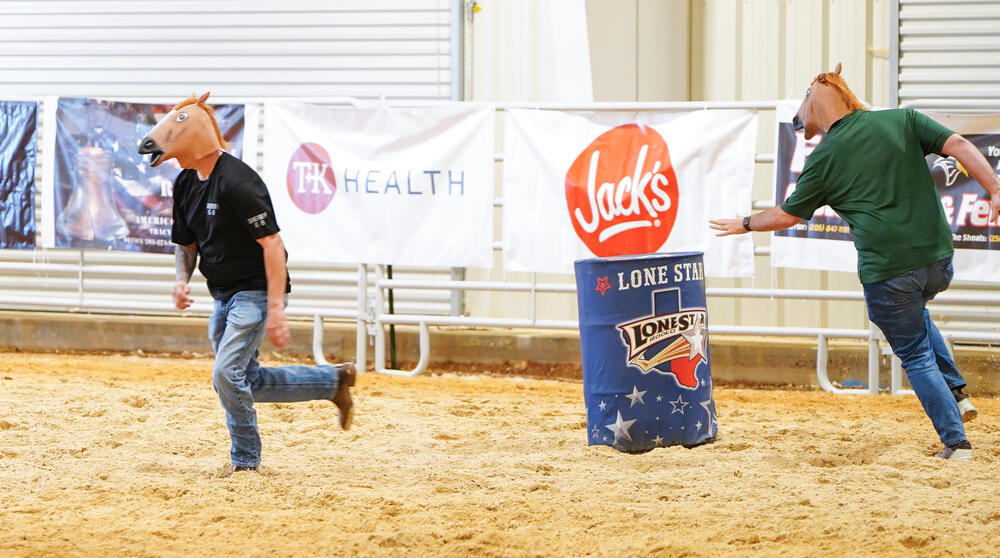 Two males with horse head masks running around a barrel in a race. 
