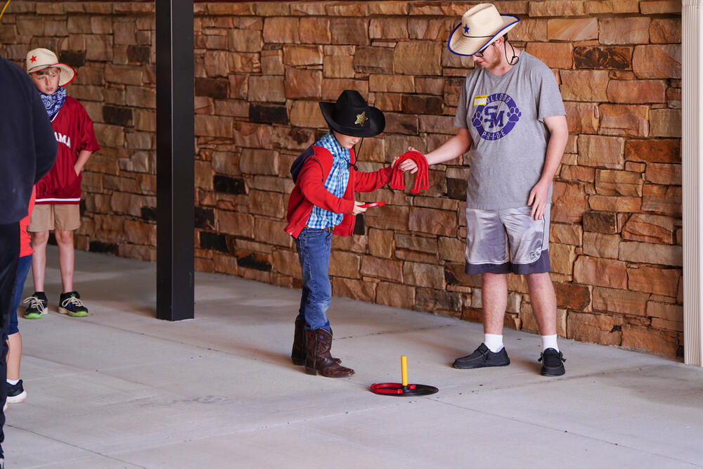 A young male dressed in western attire getting horse shoes from a volunteer to play a horse shoes game. 