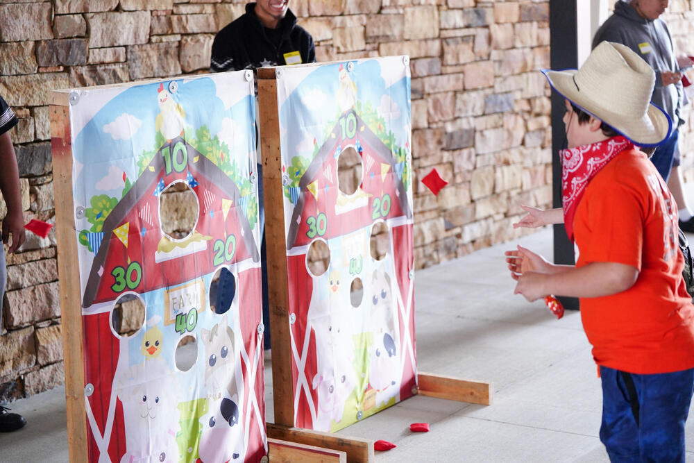 A young male wearing a cowboy hat and bandana playing a bean bag toss game. 