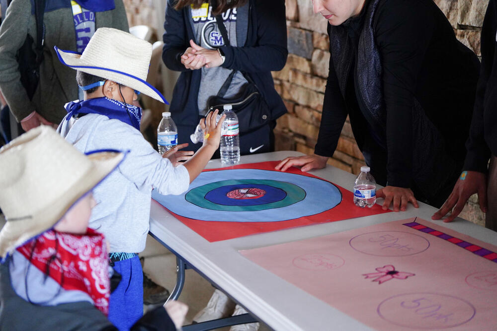 A young male wearing a cowboy hat and bandana playing a bottle flip game. 