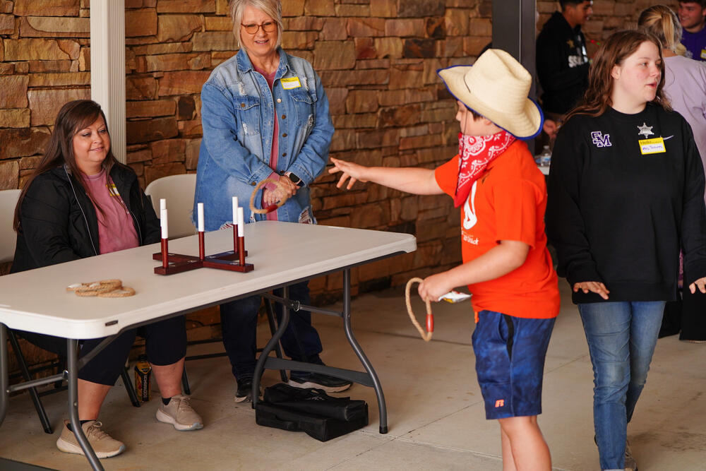 A young male wearing a cowboy hat and bandana playing a ring toss game. 